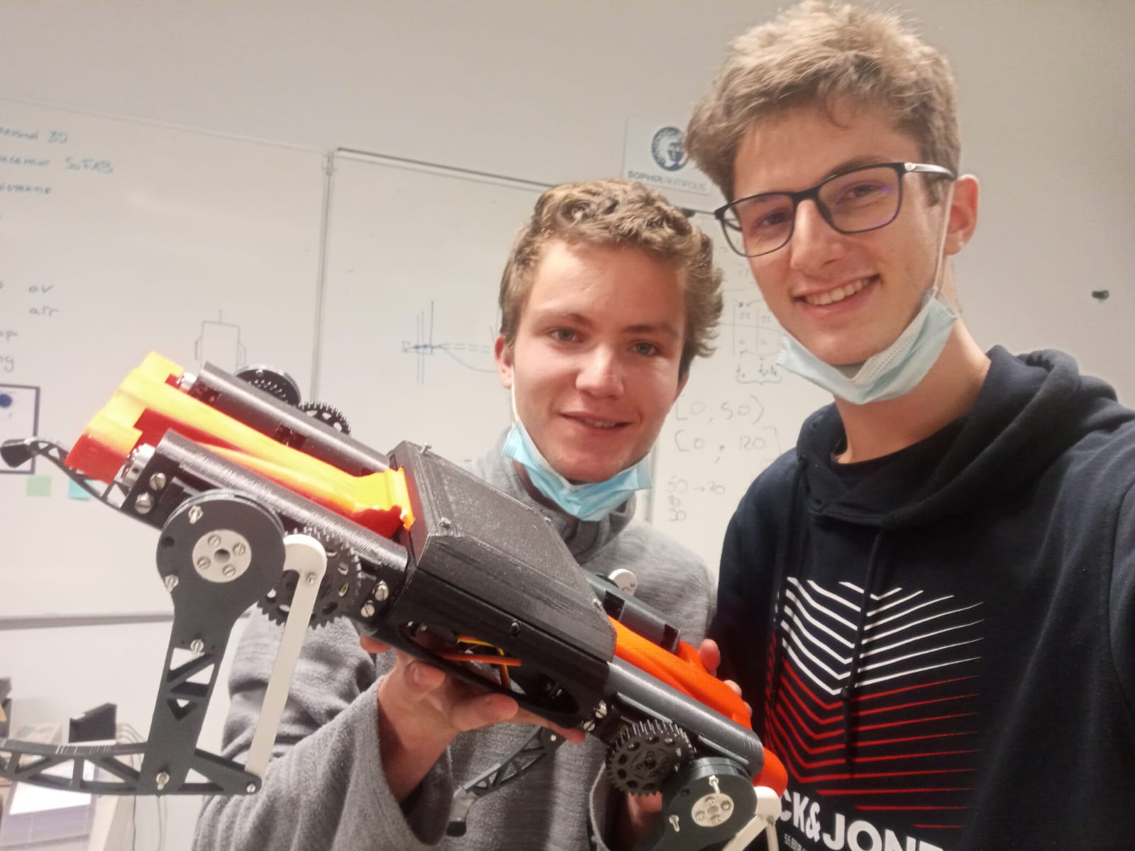 Two students smiling at the camera, masks pulled under their chins, holding up the black and orange 3D-printed robot in a classroom with a whiteboard of notes behind them.