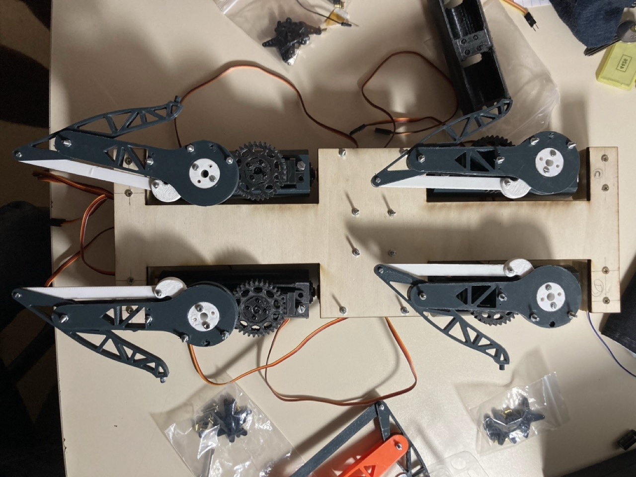The first complete v0 assembly seen from above on a workbench: four grey 3D-printed legs with visible gears and white linkage rods, bolted to a plywood plate, surrounded by bags of screws and spare parts.