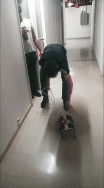 In a hallway, a person bends over the small robot holding its cable bundle, walking it forward as it takes its first steps across the tiled floor.