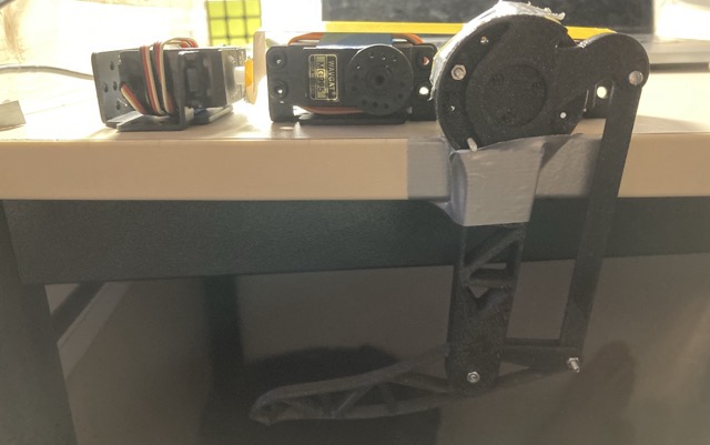 The first printed leg taped to the edge of a desk so it can hang freely, with two loose servo motors sitting on the desktop beside it.