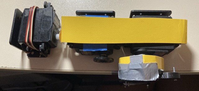 Top view of an early mock-up: two servo motors bolted to a strip of yellow-taped cardboard, standing in for the hip joint before anything was 3D printed.