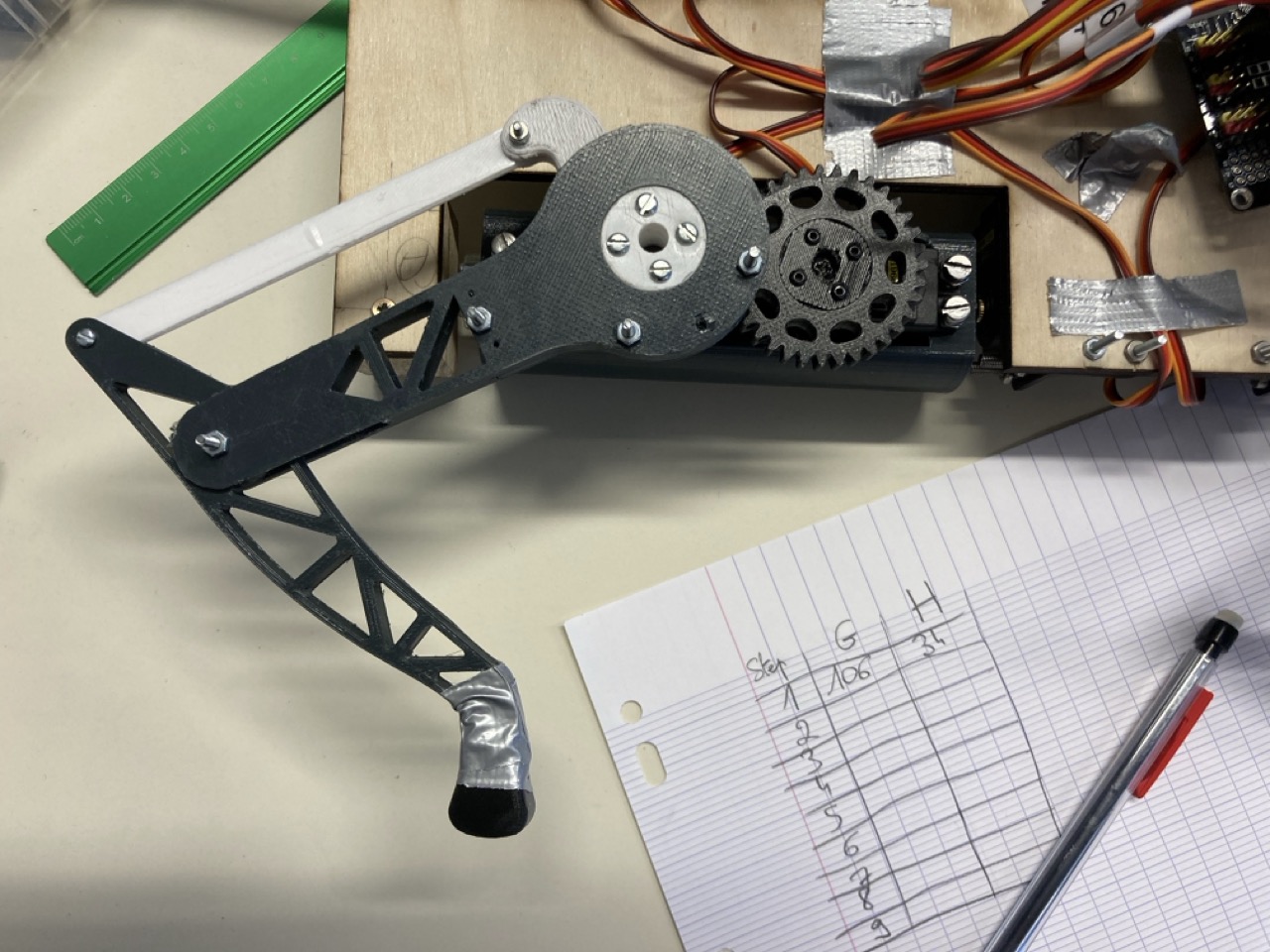 Close-up of one leg on the bench, showing the printed gear train and linkage, next to a sheet of graph paper where joint angles are being written down by hand step by step.