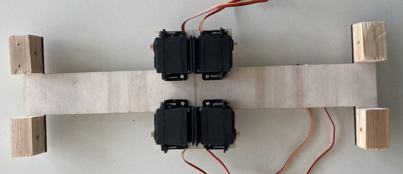 A flat plywood cross forming the first chassis, with four black servo motors bolted in pairs at the centre and wooden blocks at each corner. Servo cables trail off to the right.