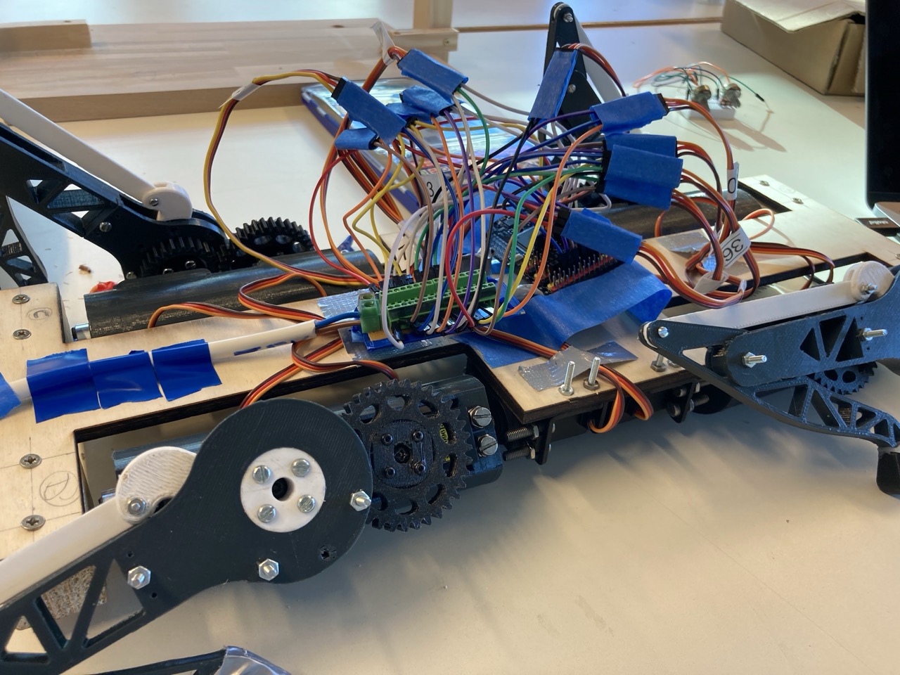 Close-up of the later v0 build: control boards buried under a dense bundle of multicoloured wires held down with blue tape, with the printed gears and legs visible around the plywood frame.