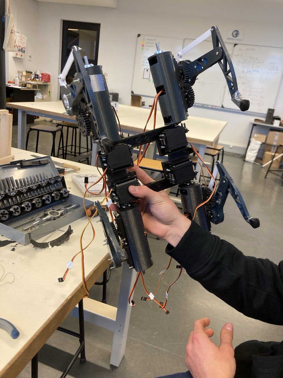 A hand holds the partly assembled v1 robot upright in a workshop, showing the black printed body, the servo motors stacked along the spine and the legs splayed out, with a tap-and-die set on the bench behind.