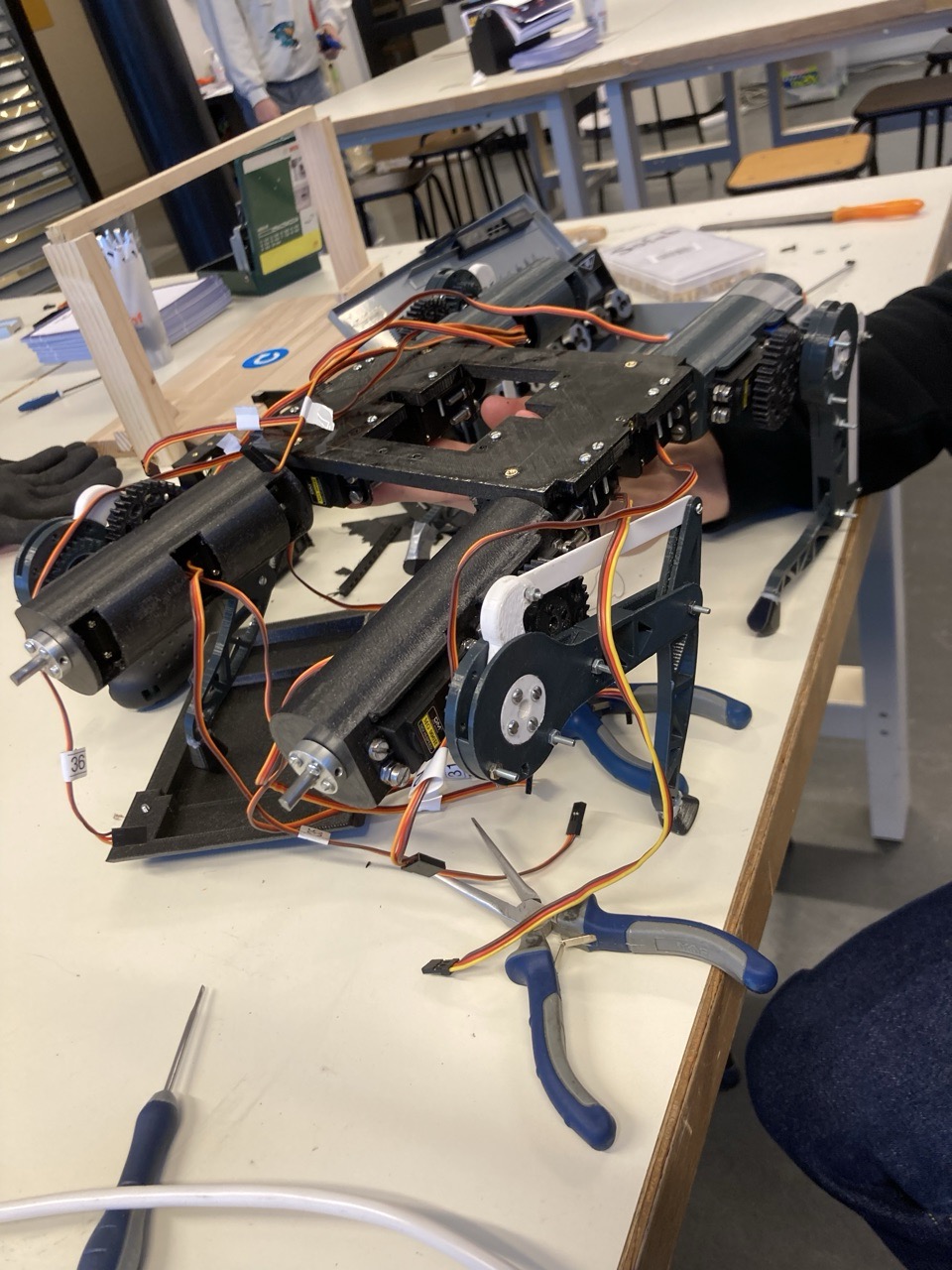 The v1 robot lying on its side on a workbench mid-assembly, orange servo cables running along the black printed frame, a pair of pliers and a screwdriver next to it.