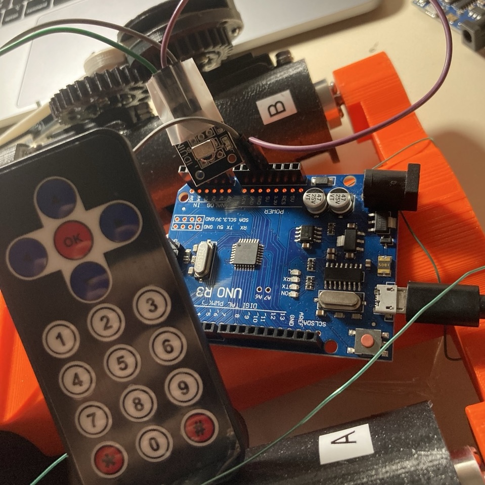 A black infrared remote control with arrow keys and a number pad, resting next to an Arduino Uno board wired into the robot's orange printed body.