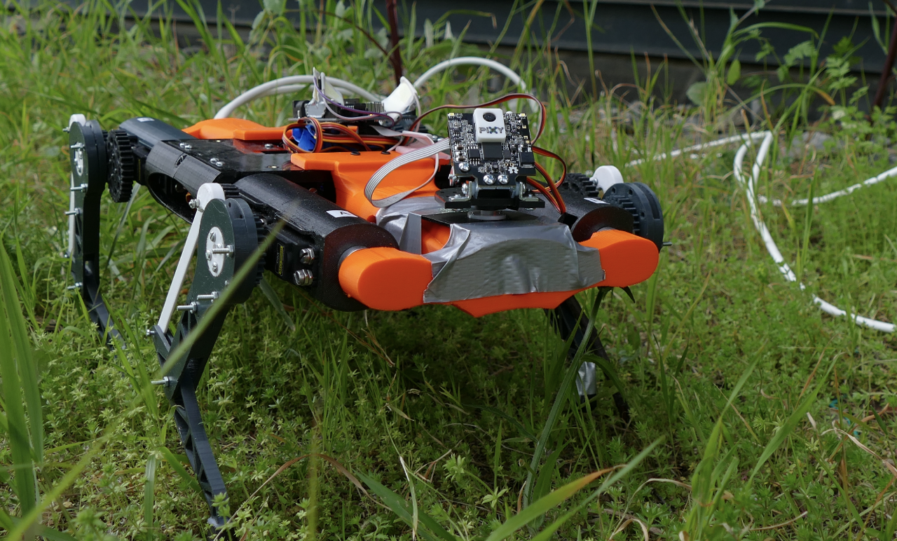 The orange and black robot standing in tall grass outdoors, a Pixy camera board mounted on the front and duct tape holding part of the shell together.