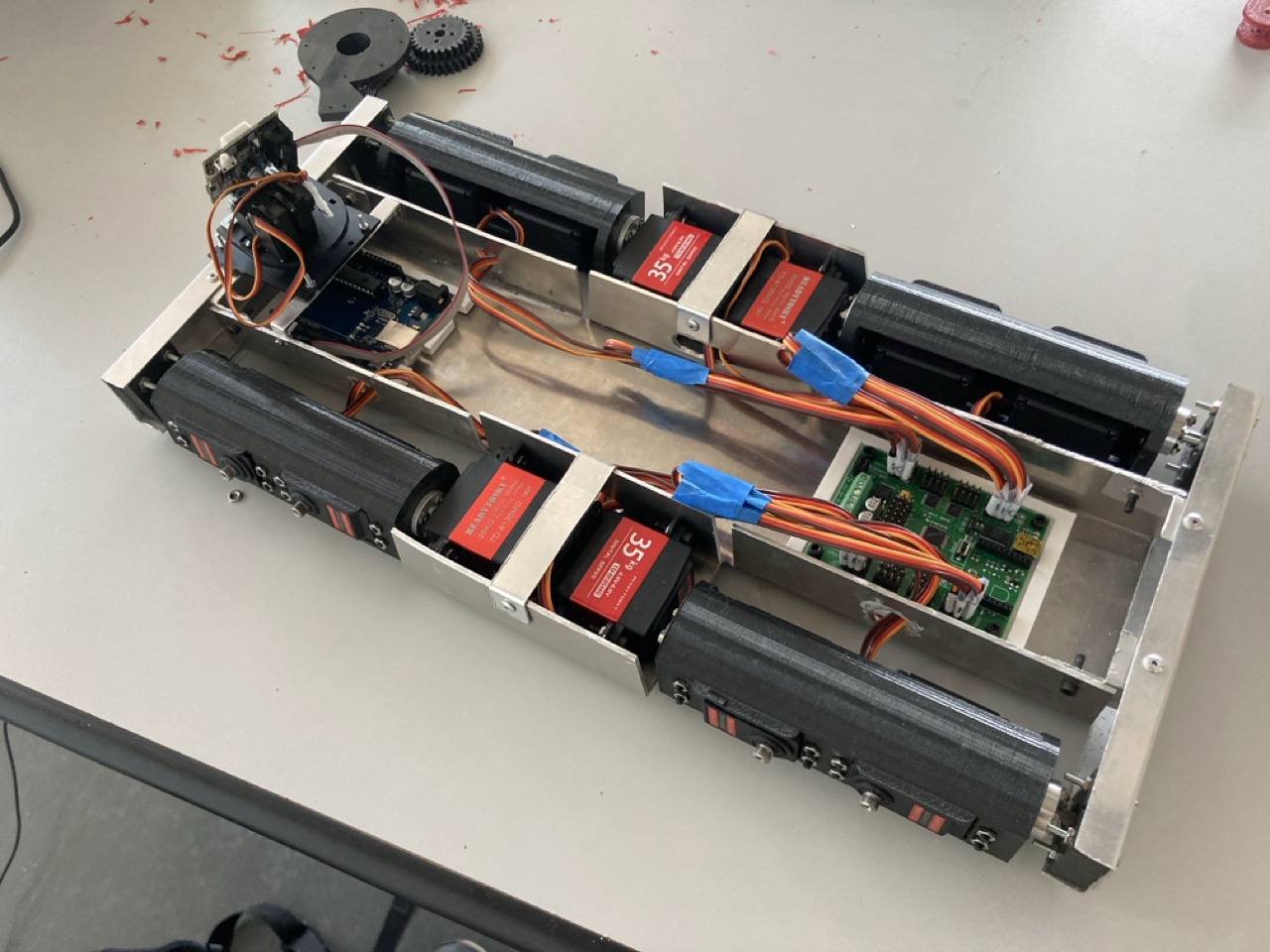 The v2 aluminium chassis on a workbench, opened up to show the motors, the red 35 kg servos strapped in place, the green controller board and the orange wiring loom running between them.