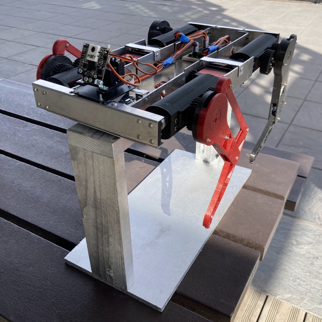 The finished robot outdoors in the sun, resting on a wooden and aluminium stand, with red 3D-printed legs, a Pixy camera mounted at the front and wiring visible along the metal frame.